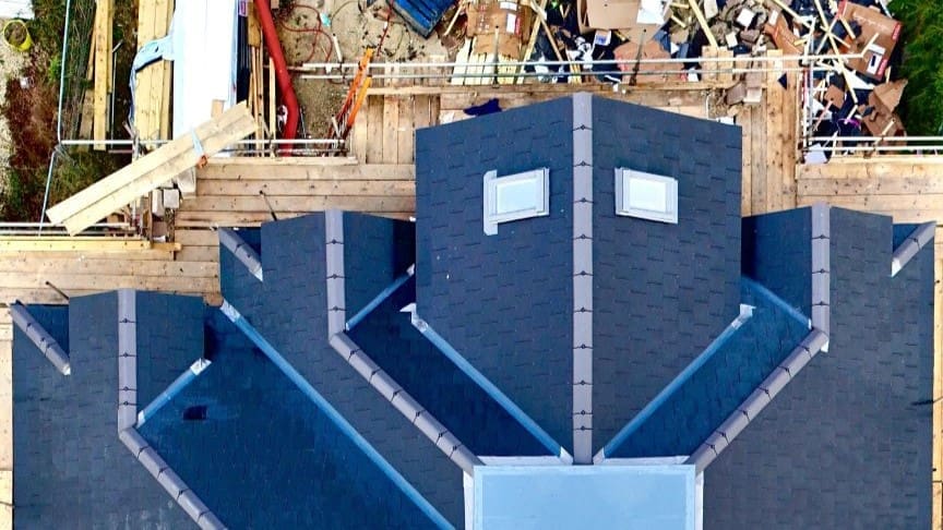 "Aerial view of a newly installed pitched roof in Broadstone, featuring dark grey slate tiles, skylights, and precise lead flashing. The roof design includes multiple ridges and valleys for effective water drainage. Surrounding scaffolding and construction materials indicate ongoing roofing work in Broadstone, highlighting expert craftsmanship in roof installation."