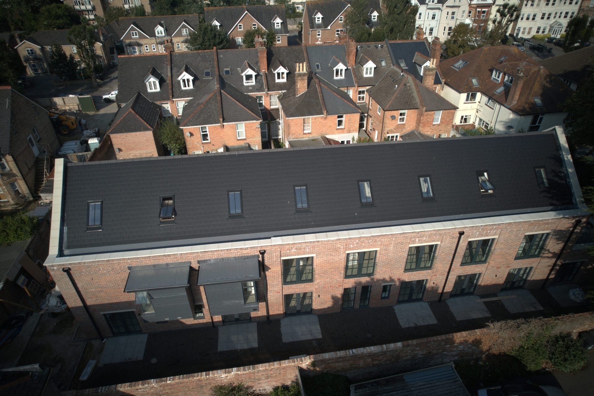 Drone shot of a contemporary brick property with a dark pitched roof featuring skylights, surrounded by traditional residential houses